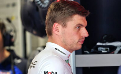 Side profile of a race car driver in a white sponsor-branded suit in a garage/pit area