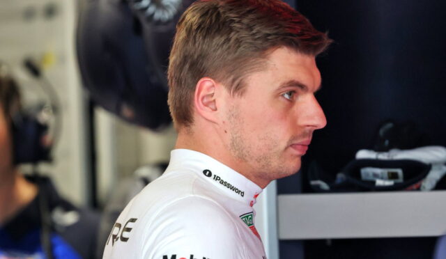 Side profile of a race car driver in a white sponsor-branded suit in a garage/pit area