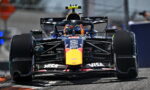 Red Bull Formula 1 car speeding toward camera on track, front wing and sponsor logos visible, driver in helmet inside cockpit with blurred pit area in background