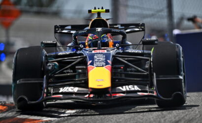 Red Bull Formula 1 car speeding toward camera on track, front wing and sponsor logos visible, driver in helmet inside cockpit with blurred pit area in background