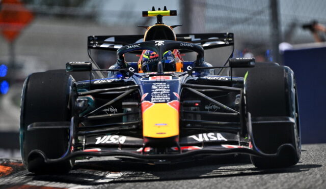 Red Bull Formula 1 car speeding toward camera on track, front wing and sponsor logos visible, driver in helmet inside cockpit with blurred pit area in background