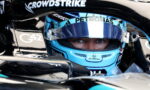 Close-up of a Formula 1 driver with a blue helmet in the cockpit, eyes visible under the visor, surrounded by sponsor logos like CrowdStrike and Petronas.