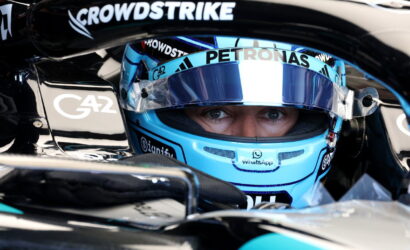 Close-up of a Formula 1 driver with a blue helmet in the cockpit, eyes visible under the visor, surrounded by sponsor logos like CrowdStrike and Petronas.