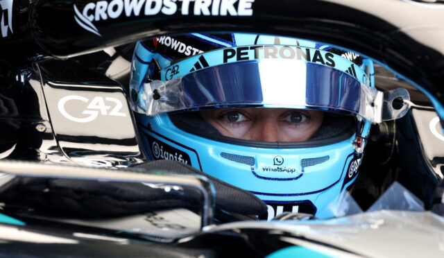 Close-up of a Formula 1 driver with a blue helmet in the cockpit, eyes visible under the visor, surrounded by sponsor logos like CrowdStrike and Petronas.