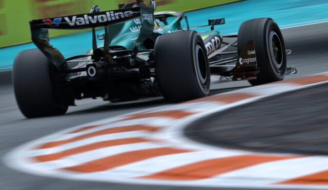 Green Formula 1 car speeds through a corner, rear view with Valvoline branding and blurred curbstones in the foreground