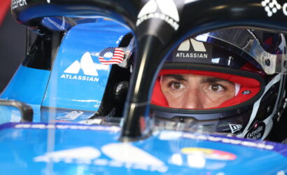Close-up of a race car driver in a red helmet, peering through the visor inside a blue Atlassian–sponsored race car