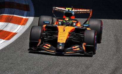 Orange Formula 1 race car speeding along a track, front view with large rear wing and slick tires on asphalt edge of a curve