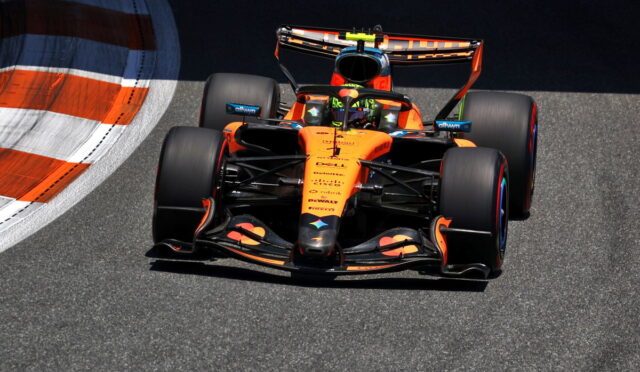 Orange Formula 1 race car speeding along a track, front view with large rear wing and slick tires on asphalt edge of a curve
