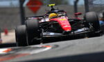 Red Formula 1 car speeding on track, driver wearing a yellow helmet, front wing close to the ground, on a blurred racing circuit backdrop with a fence in the background.