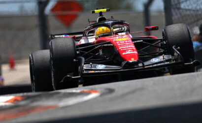 Red Formula 1 car speeding on track, driver wearing a yellow helmet, front wing close to the ground, on a blurred racing circuit backdrop with a fence in the background.