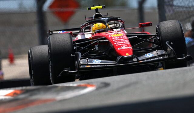 Red Formula 1 car speeding on track, driver wearing a yellow helmet, front wing close to the ground, on a blurred racing circuit backdrop with a fence in the background.