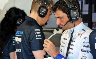 Male race car driver in white suit with sponsor logos wearing a headset, inspecting a water bottle in a pit area.
