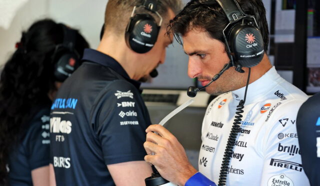 Male race car driver in white suit with sponsor logos wearing a headset, inspecting a water bottle in a pit area.