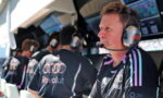 Man wearing a headset and microphone in a racing team pit, focused on the track