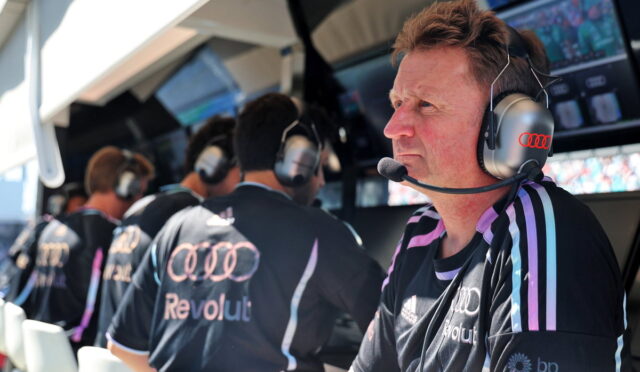Man wearing a headset and microphone in a racing team pit, focused on the track
