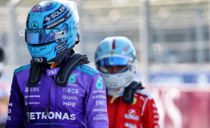 Formula 1 drivers in helmets and race suits, front driver in purple Petronas suit, back driver in red suit at pit area.