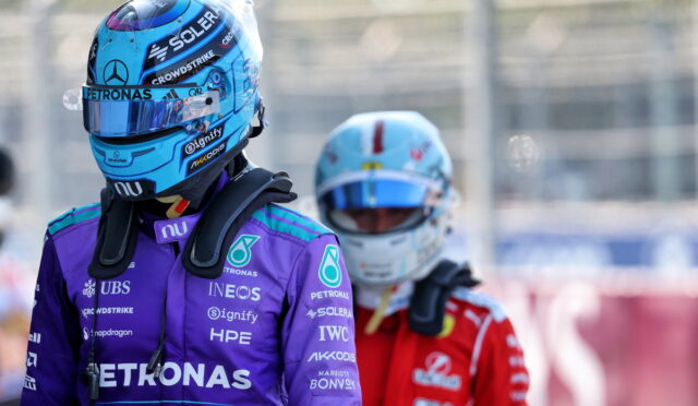 Formula 1 drivers in helmets and race suits, front driver in purple Petronas suit, back driver in red suit at pit area.