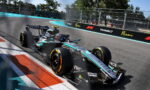 Mercedes F1 car in turquoise/black livery rounds a corner on the Miami track with sponsor barriers and fencing in the background.