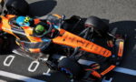 Orange Formula 1–style race car with sponsor decals sits on the track, driver wearing a neon green helmet in the cockpit.