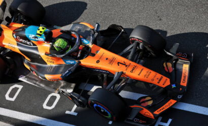 Orange Formula 1–style race car with sponsor decals sits on the track, driver wearing a neon green helmet in the cockpit.