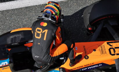 Racing driver in orange and black suit with number 81 sits in an orange Formula 1 car, wearing a colorful helmet.