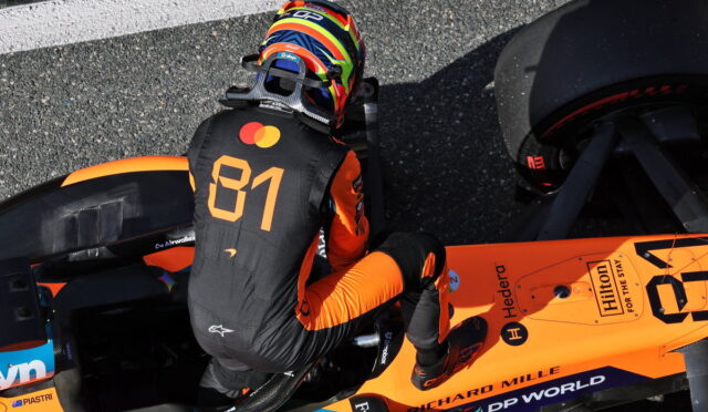 Racing driver in orange and black suit with number 81 sits in an orange Formula 1 car, wearing a colorful helmet.