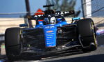 Blue Formula 1 race car on a track turning sharply, front wing and slick tires visible beneath the nose.