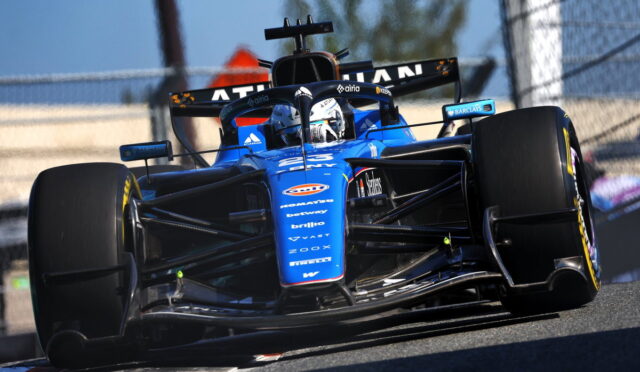 Blue Formula 1 race car on a track turning sharply, front wing and slick tires visible beneath the nose.
