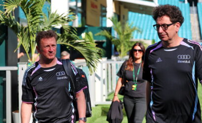 Two men in matching black athletic shirts walk through a stadium concourse, a woman in sunglasses follows behind carrying a bag and jacket.