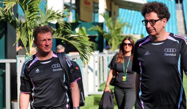 Two men in matching black athletic shirts walk through a stadium concourse, a woman in sunglasses follows behind carrying a bag and jacket.