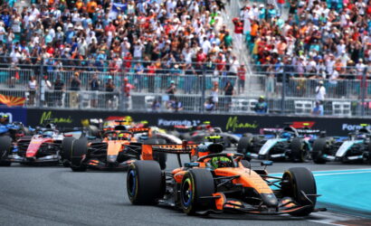 Orange Formula 1 car leads a tight pack on a race track with a large cheering crowd in the stands behind.