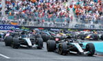 Formula 1 cars race off the start on a crowded track with spectators in the stands behind them.
