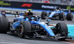 Blue Formula 1 car (Williams) on a race track, front wing close to asphalt as another car follows in the background.