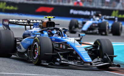 Blue Formula 1 car (Williams) on a race track, front wing close to asphalt as another car follows in the background.