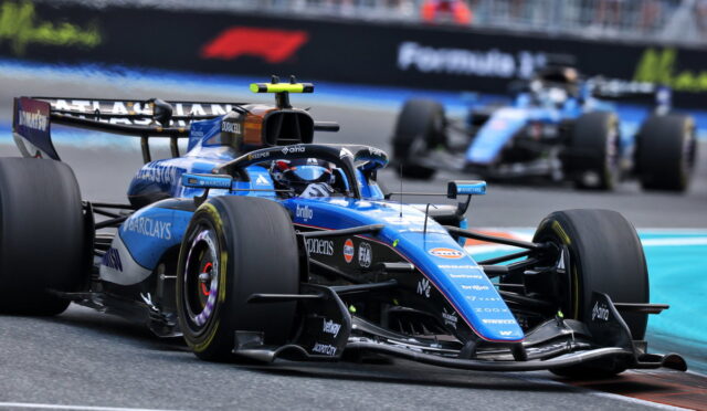 Blue Formula 1 car (Williams) on a race track, front wing close to asphalt as another car follows in the background.