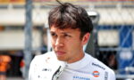 Portrait of a male race car driver in a white suit with sponsor logos, standing in a paddock behind a chain-link fence, looking pensive.