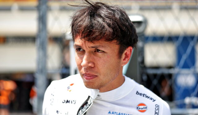 Portrait of a male race car driver in a white suit with sponsor logos, standing in a paddock behind a chain-link fence, looking pensive.