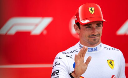 Ferrari Formula 1 driver in white racing suit waves hello against a red backdrop with F1 logos in the background.
