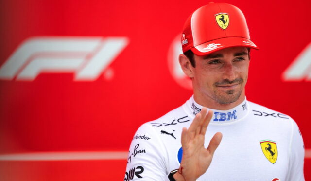 Ferrari Formula 1 driver in white racing suit waves hello against a red backdrop with F1 logos in the background.