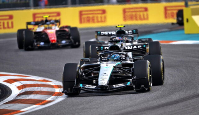 Mercedes Formula 1 car leads a pack around a bend on a race track, with yellow Pirelli barriers in the background and other F1 cars close behind.