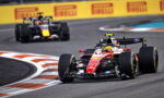Red Formula 1 car leads around a sharp chicane on a race track, close behind a pursuing black-and-yellow car, with Pirelli banners in the background.