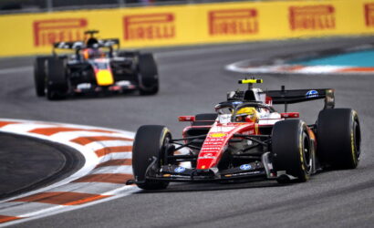 Red Formula 1 car leads around a sharp chicane on a race track, close behind a pursuing black-and-yellow car, with Pirelli banners in the background.