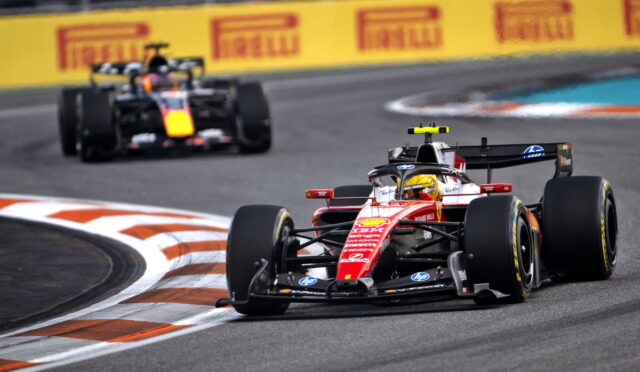 Red Formula 1 car leads around a sharp chicane on a race track, close behind a pursuing black-and-yellow car, with Pirelli banners in the background.