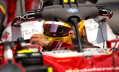 Formula 1 driver with a gold helmet inside a red single-seater, hand resting on the cockpit with sponsor logos visible (Ray‑Ban, HP).