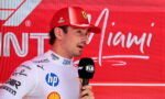 Male Formula 1 driver in white suit giving interview, holding a FIA microphone with a red sponsor backdrop behind him.