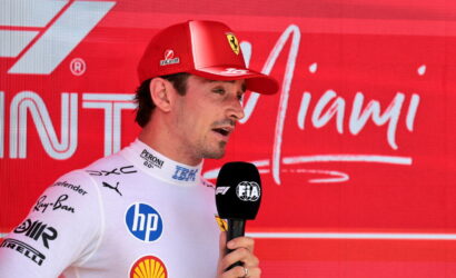 Male Formula 1 driver in white suit giving interview, holding a FIA microphone with a red sponsor backdrop behind him.