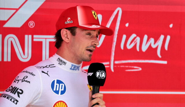 Male Formula 1 driver in white suit giving interview, holding a FIA microphone with a red sponsor backdrop behind him.