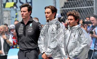 Three Mercedes team members stand in the paddock, two wearing silver jackets and one in a black polo, with spectators in the background.