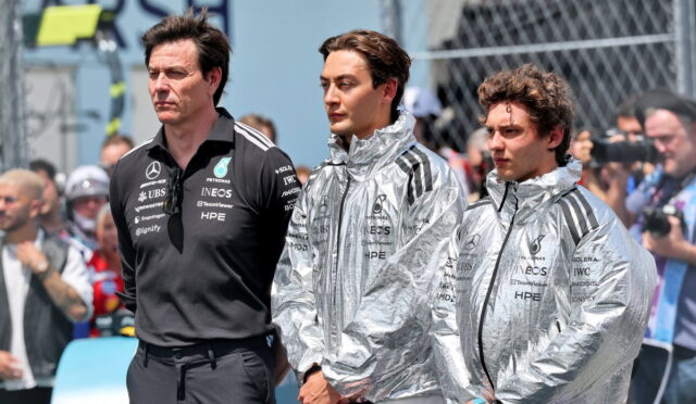 Three Mercedes team members stand in the paddock, two wearing silver jackets and one in a black polo, with spectators in the background.