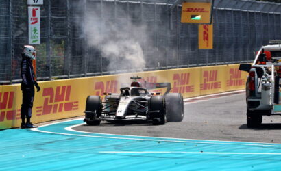 Formula 1 car exiting pit lane with smoke from tires as a crew member watches from the track edge, DHL banners along the barrier.